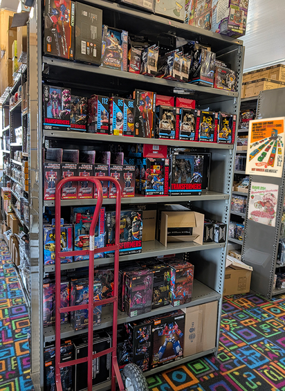 Metal shelving unit stocked with boxed Transformers action figures and toys beside a red hand truck on colorful geometric carpet
