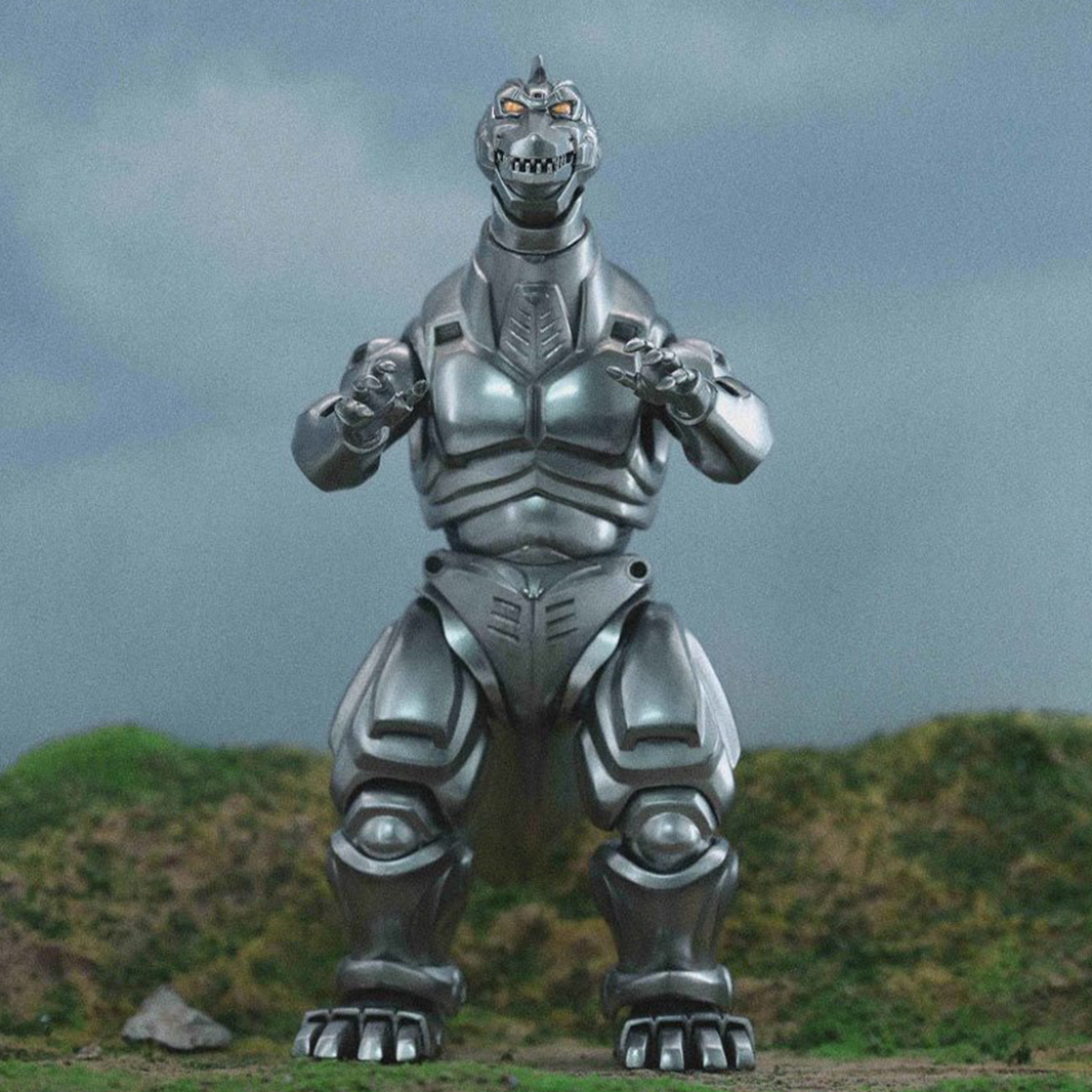 Posed metallic robot dinosaur figure with glowing eyes standing on grassy terrain against a cloudy sky background.