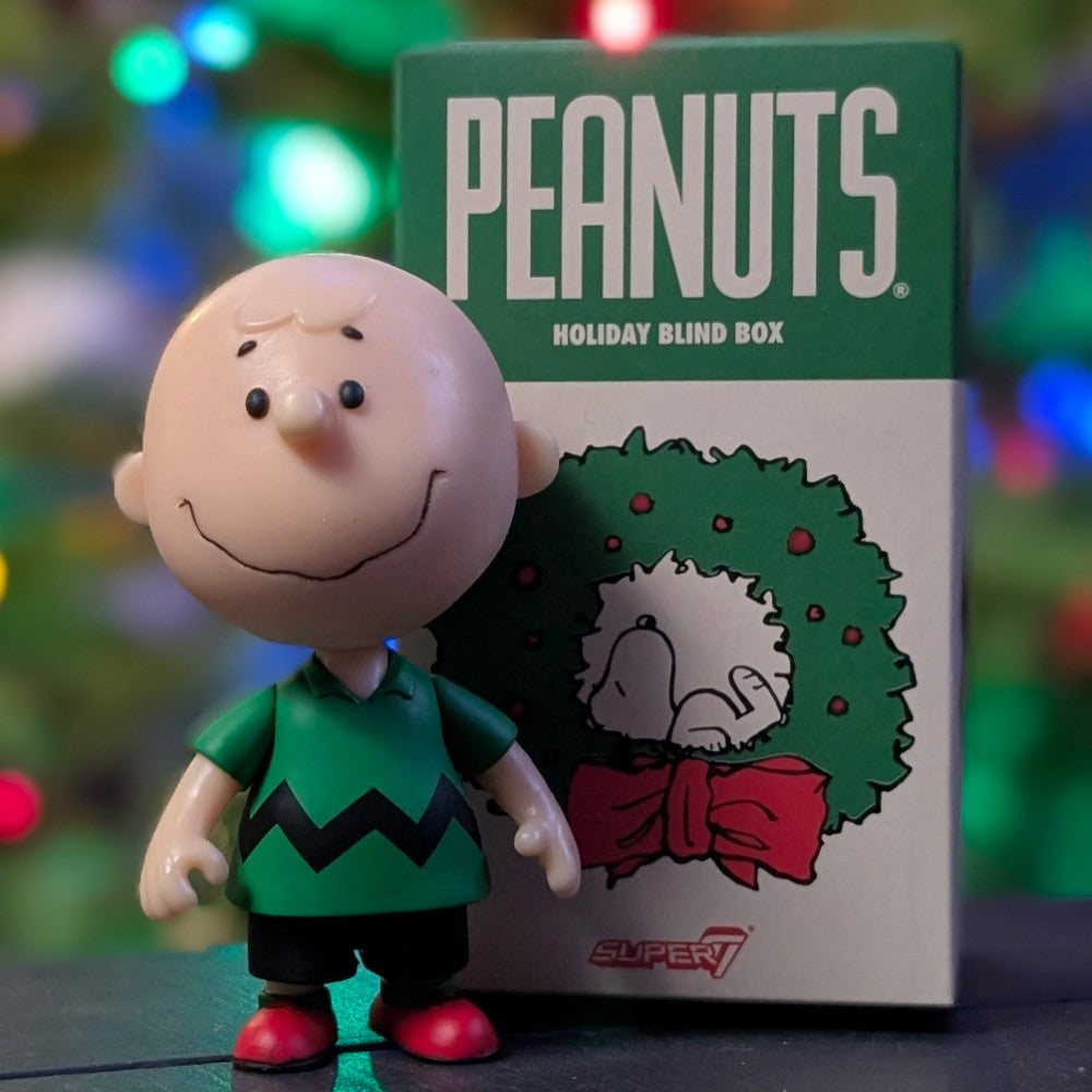 Loose Super7 Charlie Brown figure in green shirt with black zigzag standing next to Peanuts Holiday Blind Box with wreath design