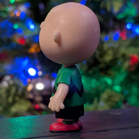 Loose Super7 Charlie Brown figure in green zigzag shirt standing in profile with colorful holiday lights blurred behind him
