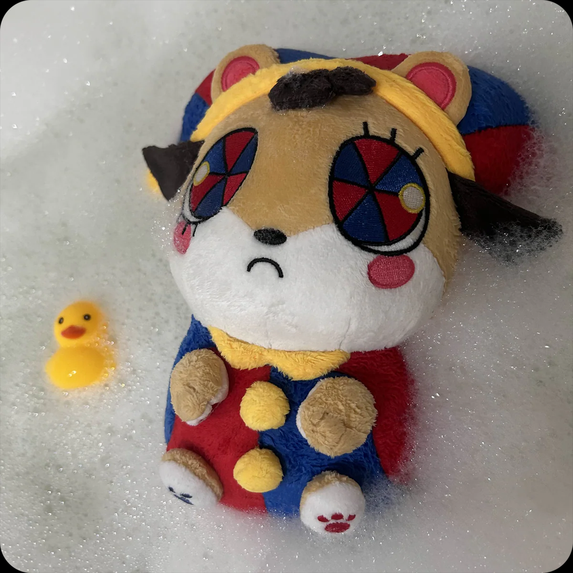 Plush character with patterned eyes and colorful outfit lying in foam bubbles next to a small yellow rubber duck.