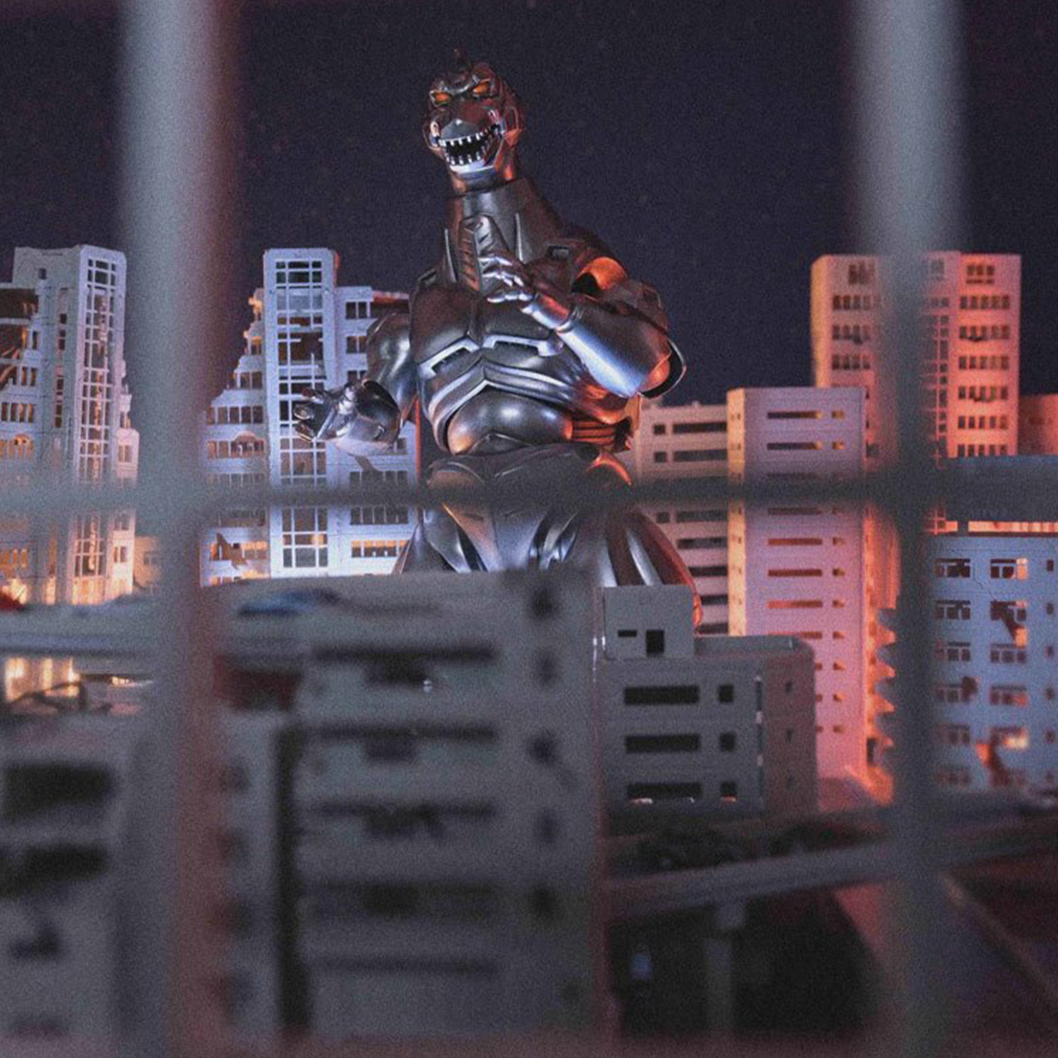 Metallic robot dinosaur figure posed amid miniature city buildings illuminated by warm night lighting.