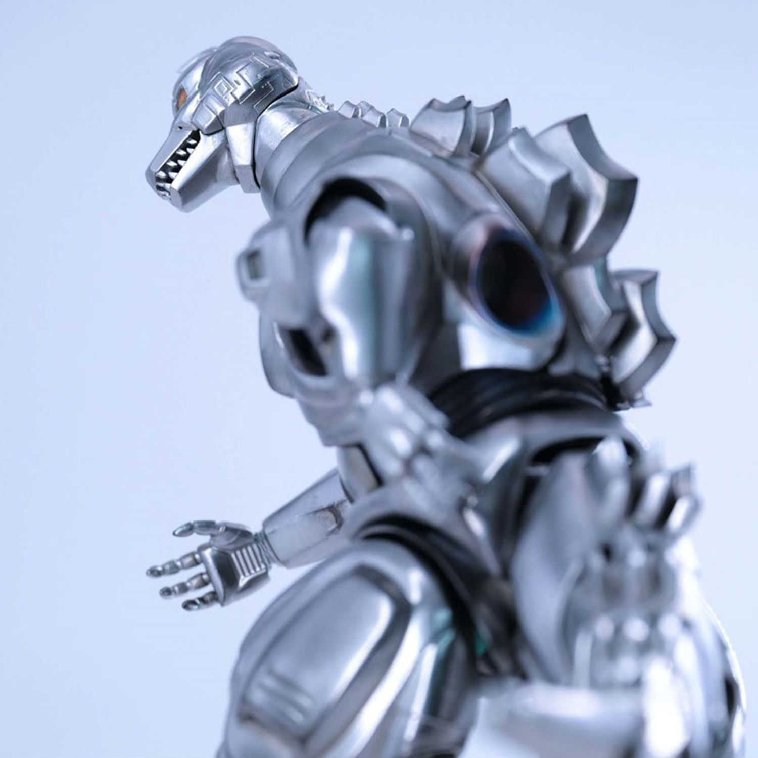 Close-up of a silver mechanical dinosaur figure with articulated hands and segmented armor against a pale blue background