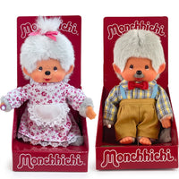 Two Monchhichi dolls seated in red display holders, one wearing floral dress, the other in plaid shirt and overalls.