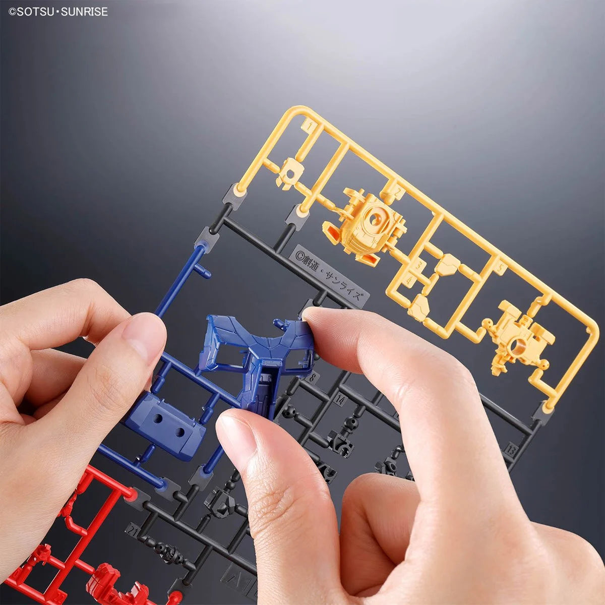 Hands assembling blue and yellow plastic model parts from a sprue frame against a gray gradient background