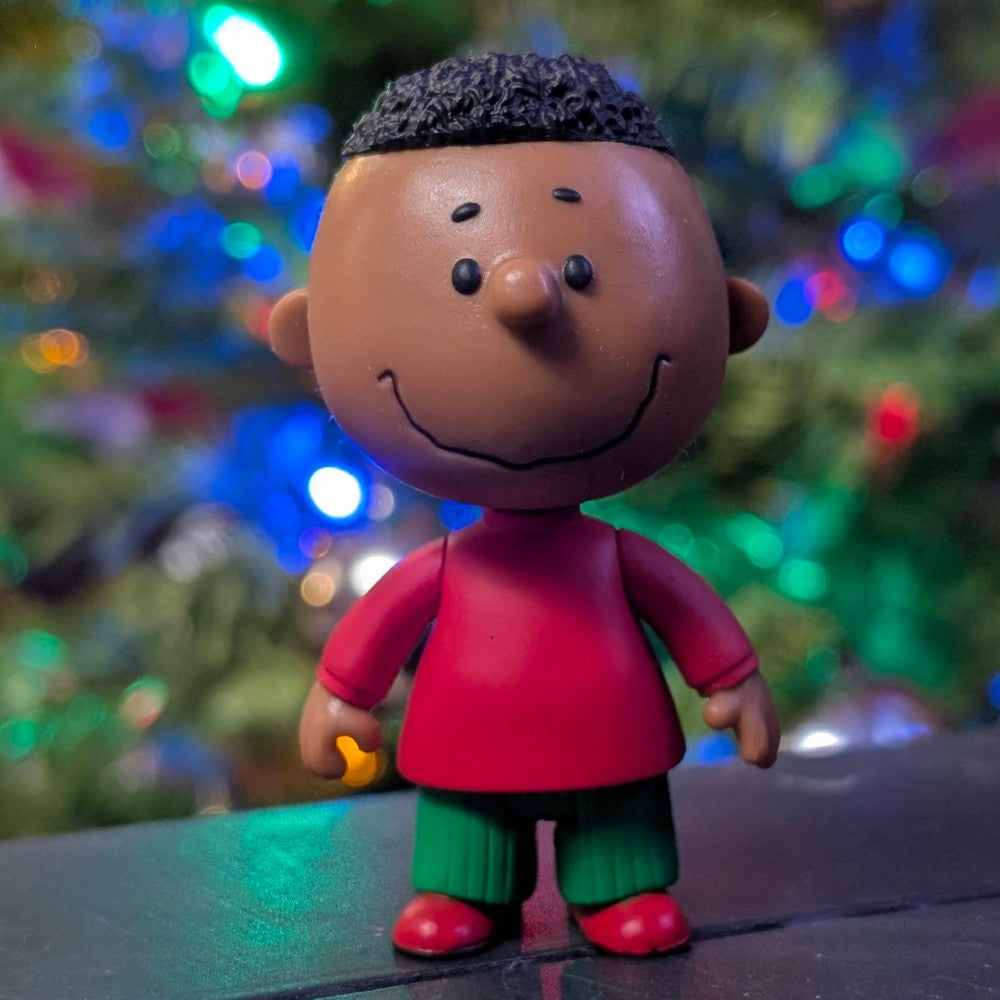 Super7 Franklin figure in red shirt and green pants standing with colorful holiday lights softly blurred behind
