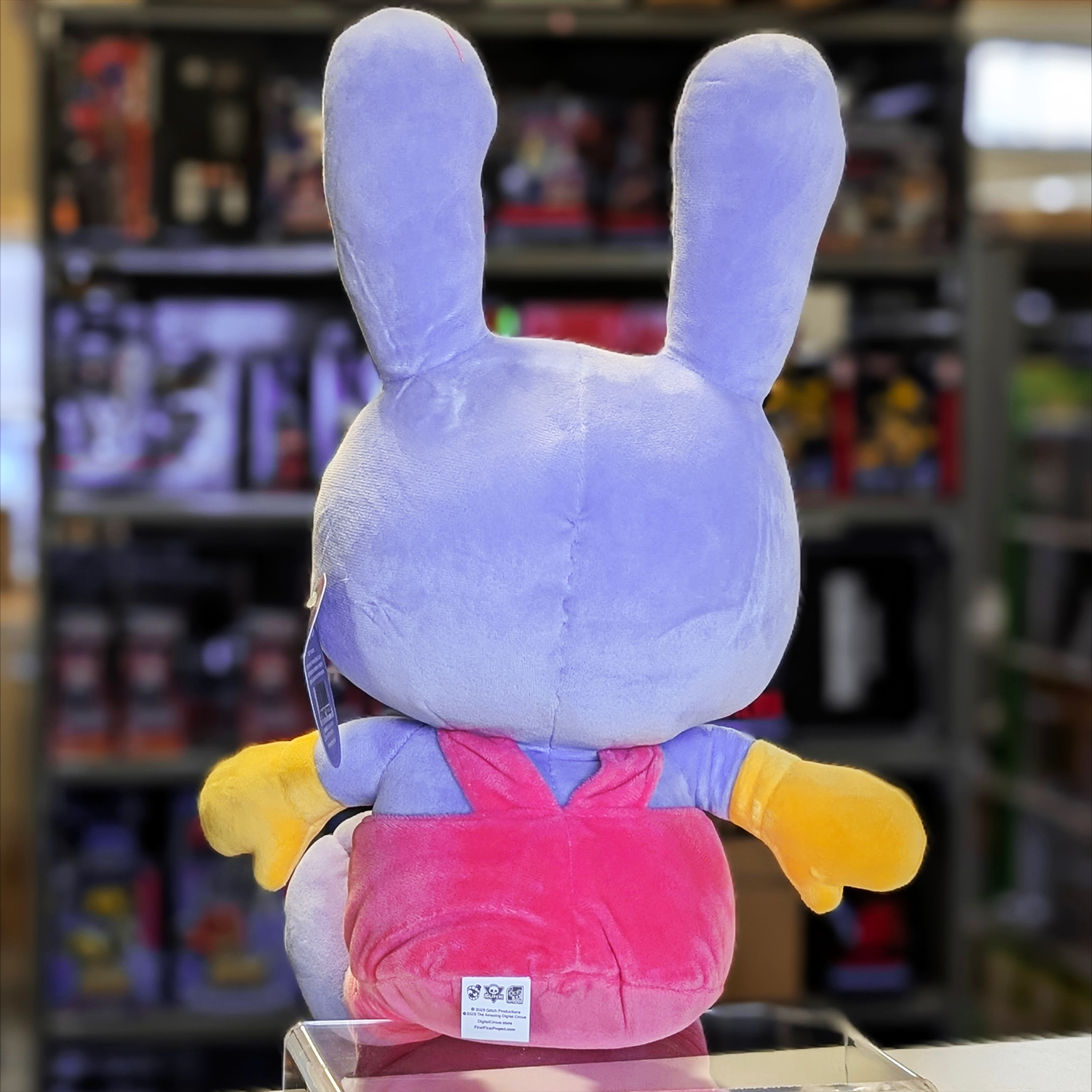 Rear view of Glitch Jax plush toy in purple with pink overalls and yellow gloves from The Amazing Digital Circus collection