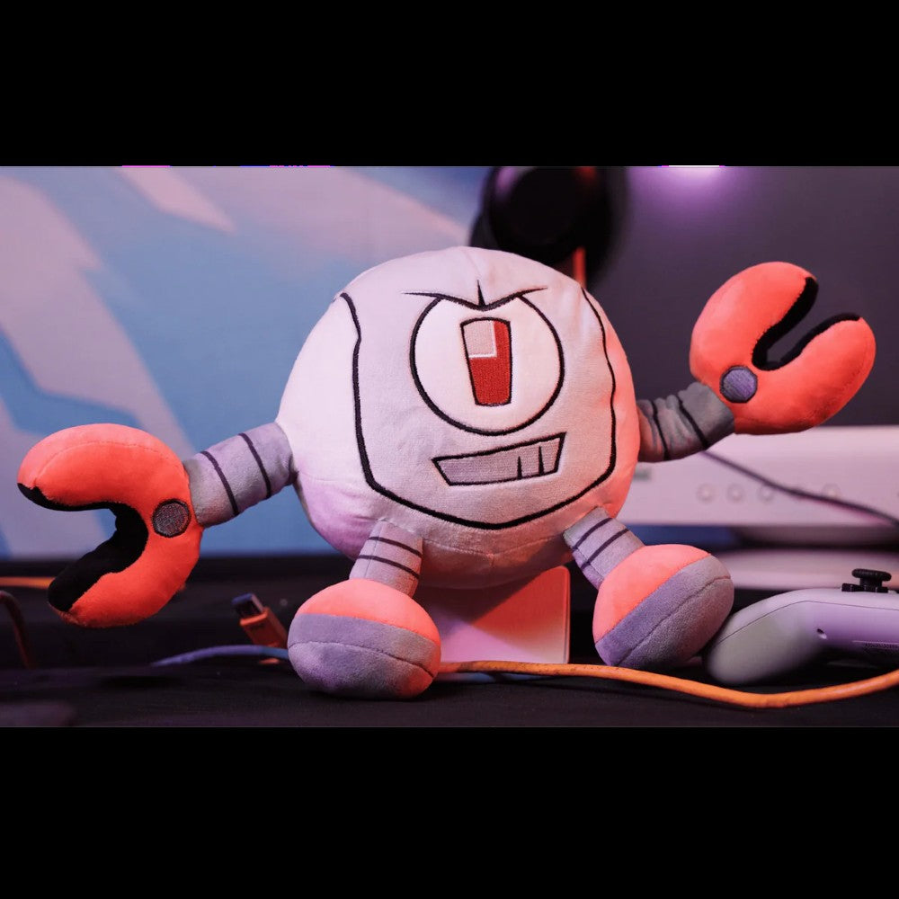 Kaboodle Plush Gameoverse Official Collectible Figure by Glitch Productions with gray body and orange clawed arms on desk next to controller