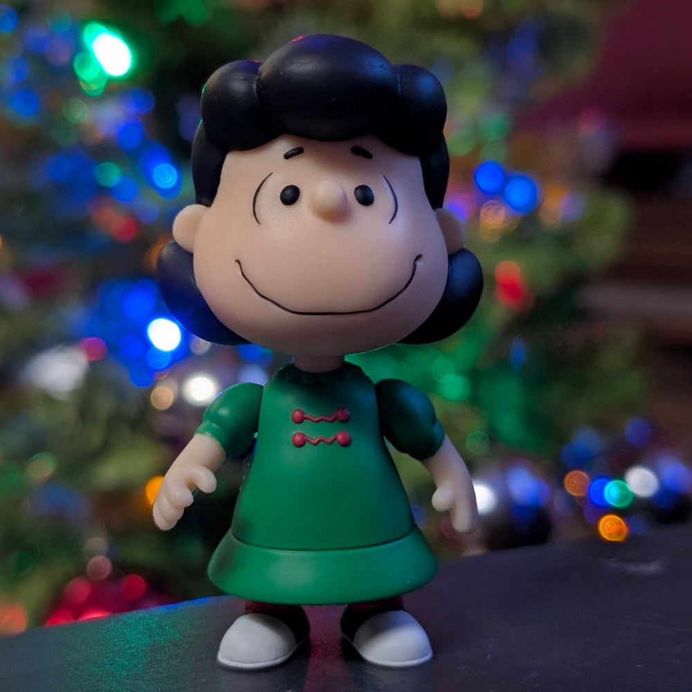 Super7 Lucy Van Pelt ReAction figure in holiday green dress with smiling expression and festive tree lights in background