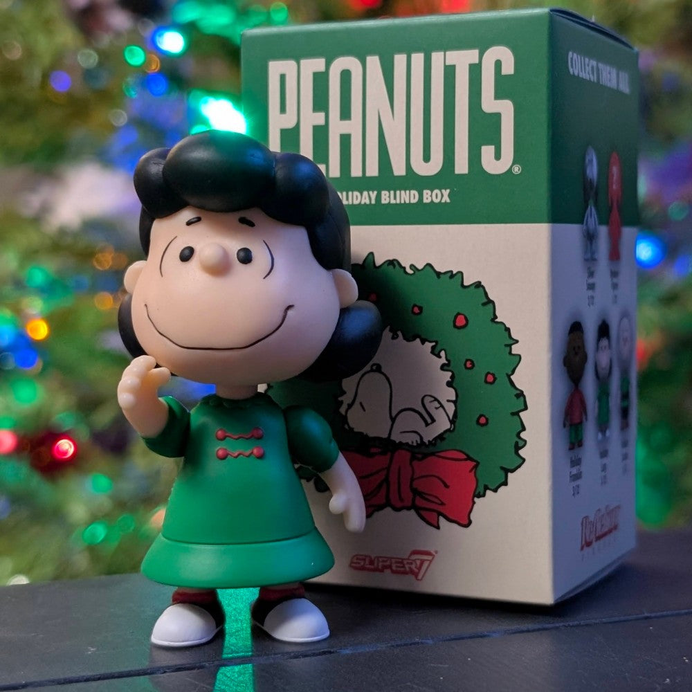 Loose Lucy VanPelt figure in green dress standing in front of Super7 Peanuts Holiday Blind Box with Christmas tree lights background
