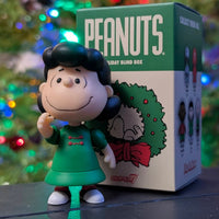Loose Lucy VanPelt figure in green dress standing in front of Super7 Peanuts Holiday Blind Box with Christmas tree lights background