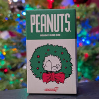 Super7 Peanuts Holiday Blind Box with Snoopy inside festive green wreath and red bow against colorful holiday lights background