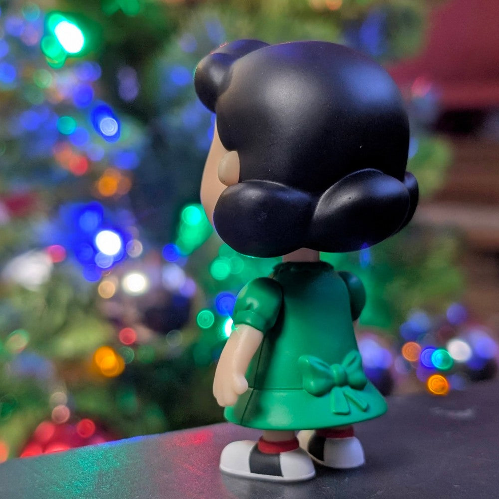 Loose Super7 Lucy Van Pelt figure in green dress with bow, shown from the back against colorful holiday lights background