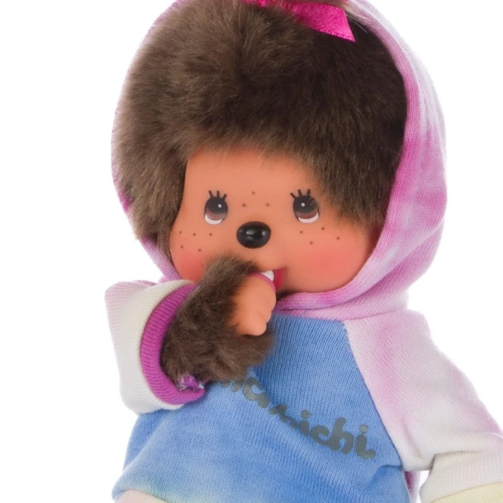 A close-up of a Chocochip doll with brown faux fur hair, a pink bow, and a blue hooded sweatshirt with pink and white tie-dye details, set against a plain white background.