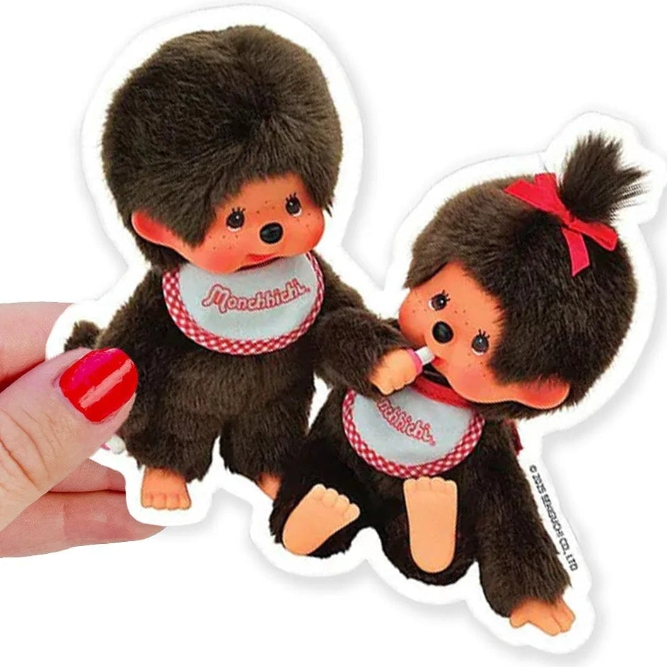 Two small plush toys resembling a monkey with a red bow and a heart-shaped tag, held by a hand with red nail polish.
