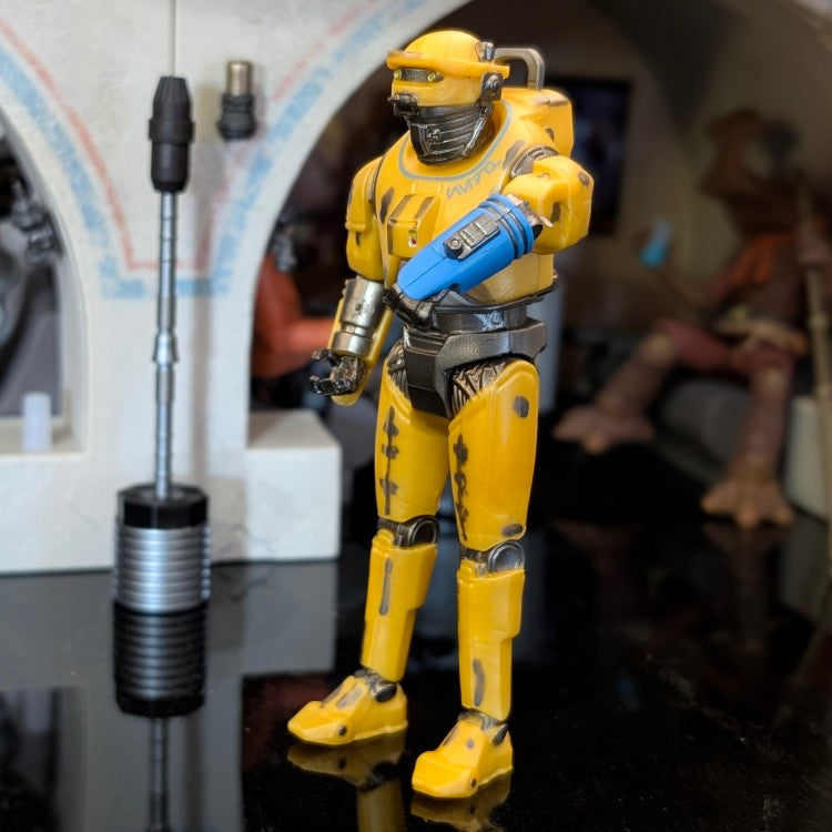 Hasbro NED-B Star Wars 3.75 inch Droid Factory action figure in yellow with blue arm detail on display.