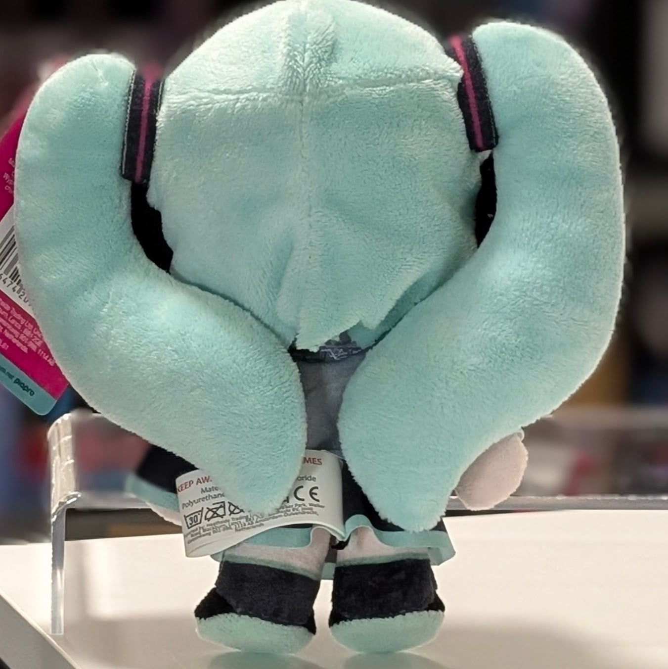 Rear view of UCC branded 6-inch Hatsune Miku plush with twin teal ponytails and care label displayed on a white surface