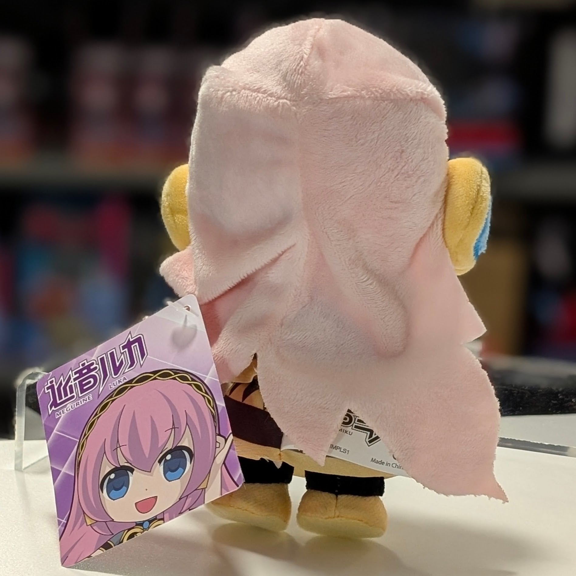 UCC 6-inch Megurine Luka plush viewed from the back with pink hair and yellow headphones holding a tag