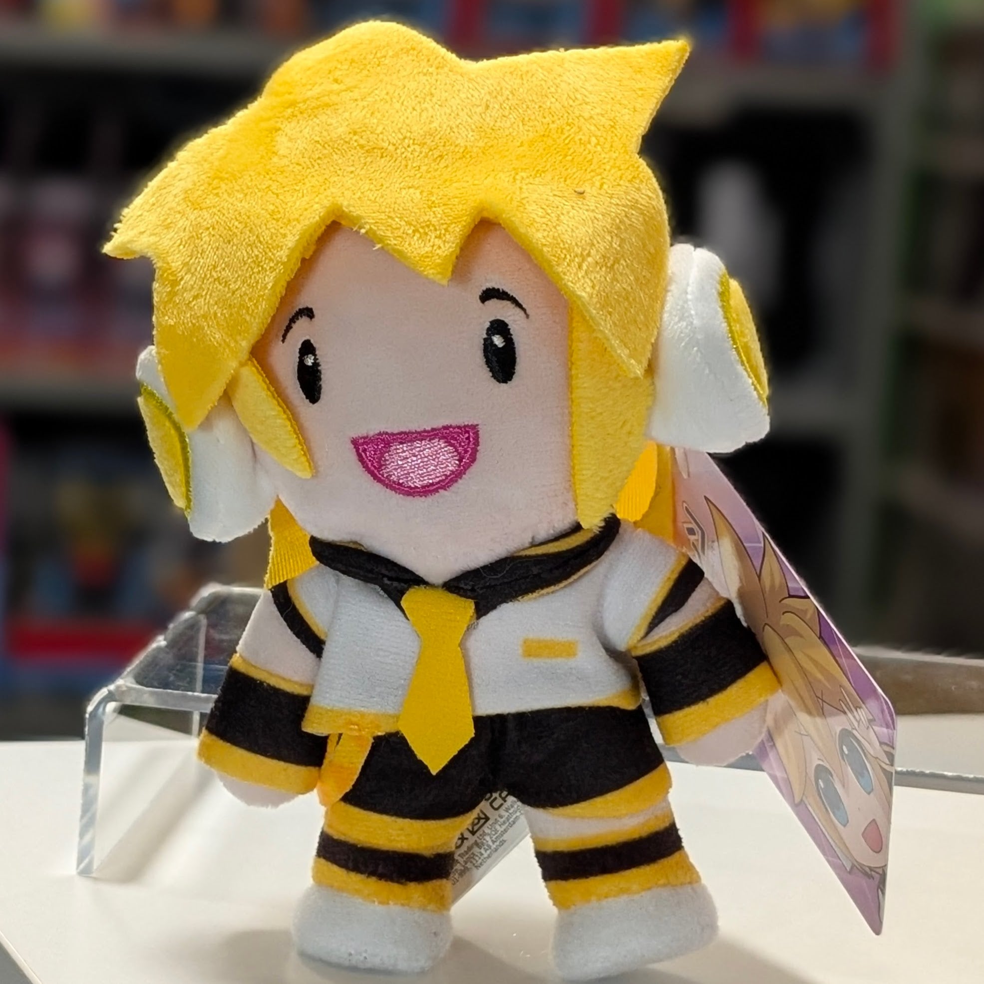 UCC 6-inch Kagamine Len plush with bright yellow hair, black and yellow outfit, smiling with product tag on blurred store background