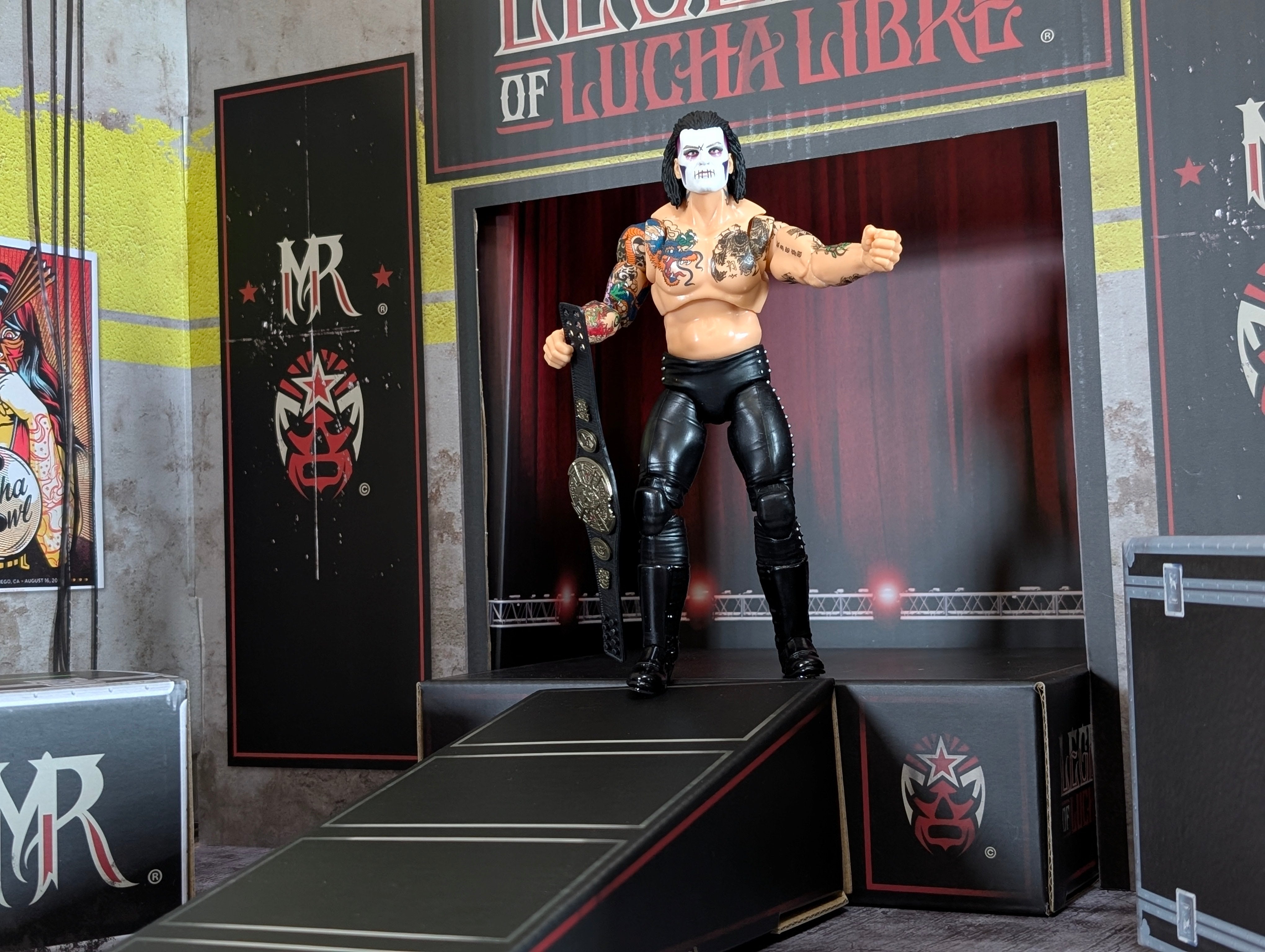 Action figure wrestler with face paint and tattoos holding a championship belt on a stage ramp with Lucha Libre branding.