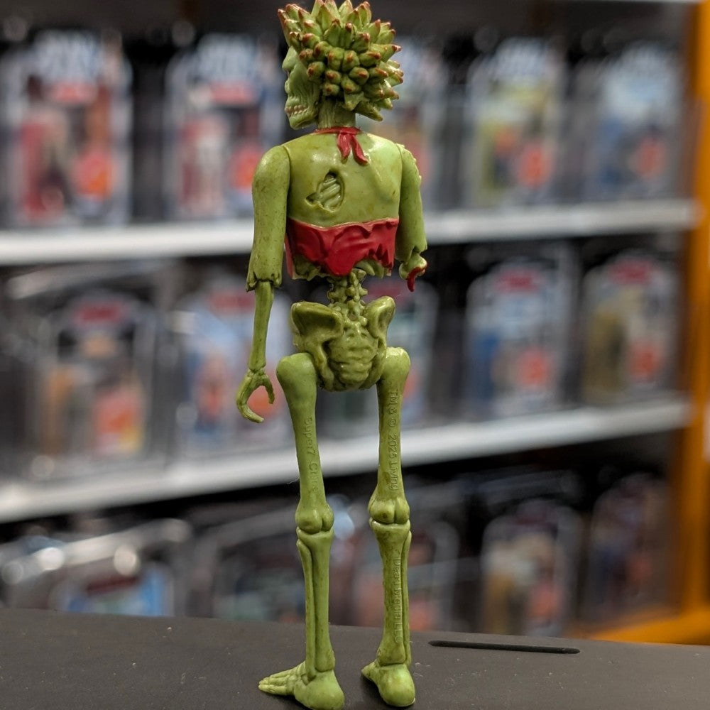Super7 “Trash” neon green female zombie figure with torn red top shown from behind on blurred store shelf background