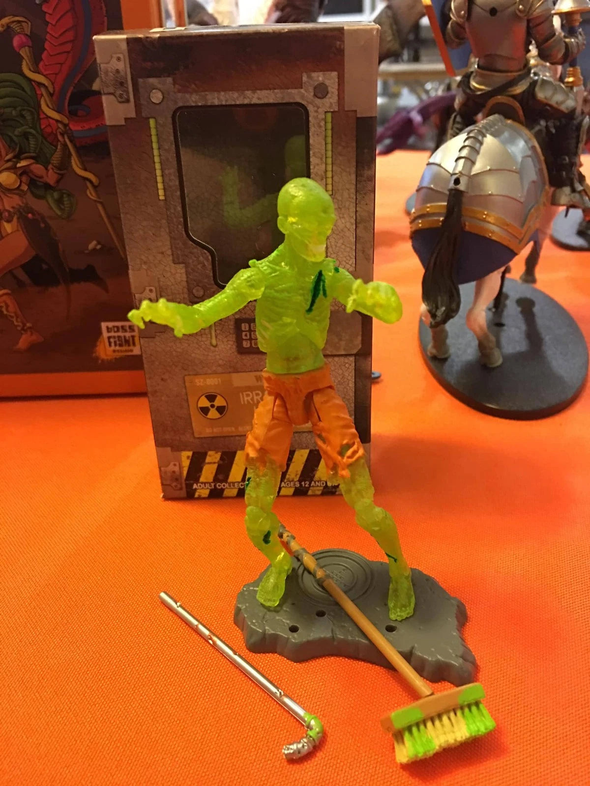 Toy figure of a character with a broom and brush on an orange surface, with packaging in the background.