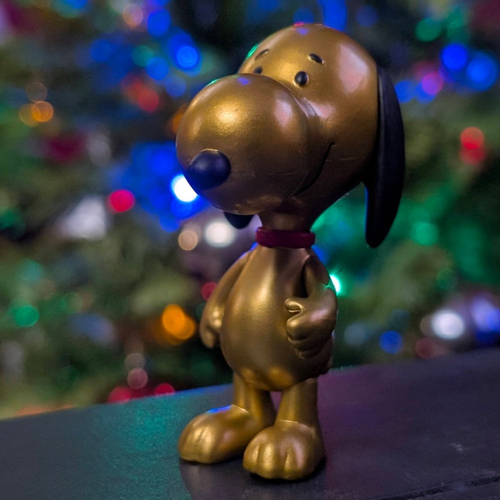 Shiny gold metallic Snoopy figure by Super7 with black ears and red collar against blurred colorful holiday lights background