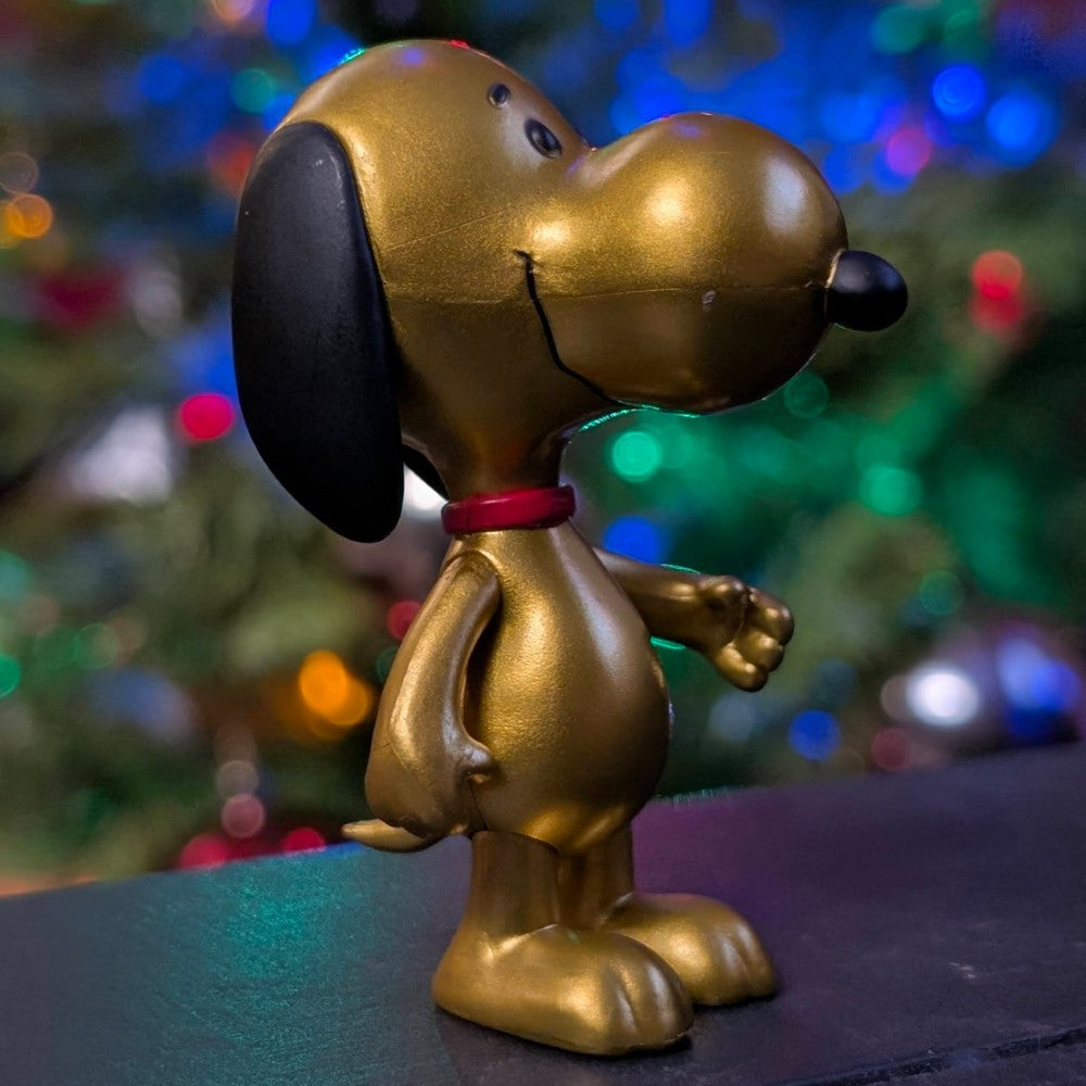 Shiny golden Snoopy figure by Super7 with black ears and red collar against colorful holiday light background