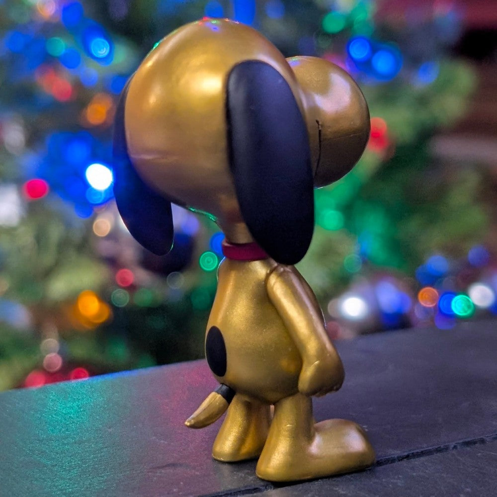 Loose Super7 shiny gold Snoopy figure with black ears and red collar against blurred colorful holiday lights background