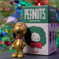Golden metallic Snoopy figure by Super7 standing next to Peanuts holiday blind box with festive blurred tree lights background