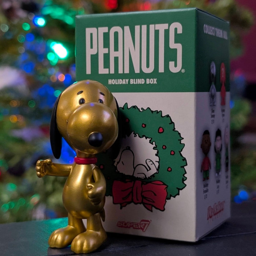 Gold metallic Snoopy figure from Super7 Holiday Blind Box standing beside Peanuts themed packaging and festive background lights