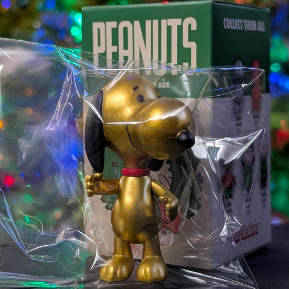 Shiny golden Snoopy figure by Super7 standing in front of Peanuts holiday blind box with festive background lights
