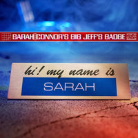 Metal nameplate reading "hi! my name is SARAH" with a blurred blue and red background from Doctor Collector